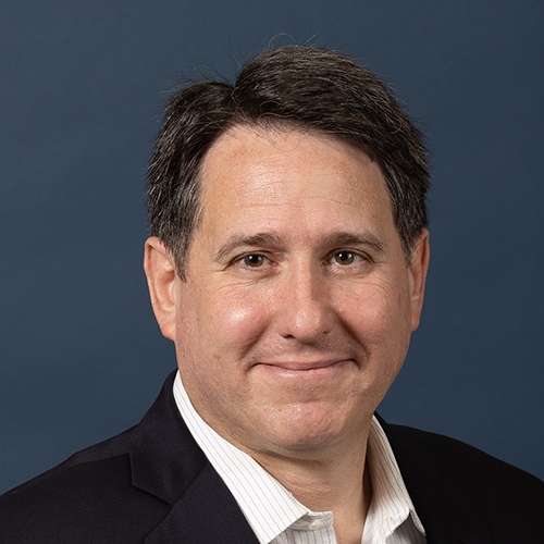 A man in a suit and white shirt smiles against a plain blue background.